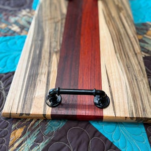 May include: A rectangular wooden serving tray featuring a central dark red and brown wood panel flanked by lighter wood with dark grain patterns. The tray includes two black industrial-style metal pipe handles attached at each end.