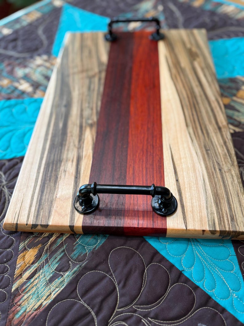 May include: A rectangular wooden serving tray featuring a central dark red and brown wood panel flanked by lighter wood with dark grain patterns. The tray includes two black industrial-style metal pipe handles attached at each end.