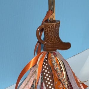 May include: A brown ceramic cowboy boot ornament with a decorative ribbon skirt. The boot has embossed details and is attached to a wooden clip. The skirt is made of various patterned ribbons in shades of orange, brown, and cream.