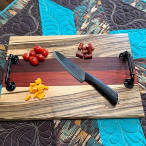 May include: A rectangular wooden serving board featuring horizontal stripes of light and dark wood grain, equipped with two black industrial-style metal pipe handles. A black chef knife, red cherry tomatoes, small sausage pieces, and yellow cheese cubes.
