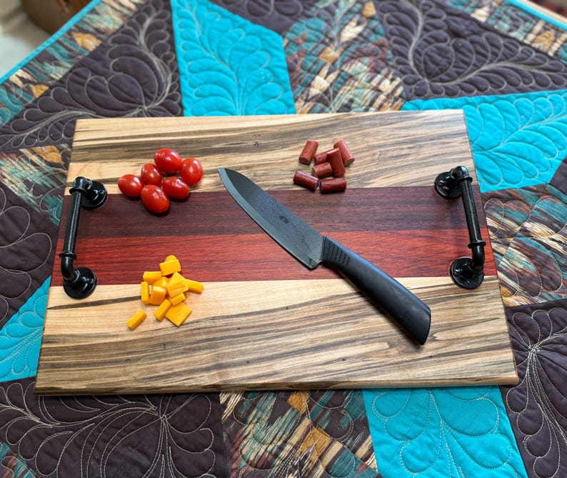 May include: A rectangular wooden serving board featuring horizontal stripes of light and dark wood grain, equipped with two black industrial-style metal pipe handles. A black chef knife, red cherry tomatoes, small sausage pieces, and yellow cheese cubes.