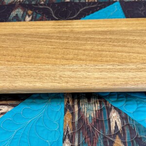 May include: A rectangular wooden cutting board with a light brown finish. The board has a smooth surface and rounded edges. It is placed on a patterned quilt with teal and dark blue colors.
