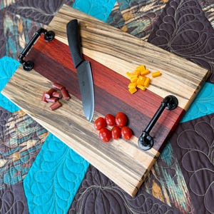 May include: A rectangular wooden serving board featuring alternating stripes of light wood with dark grain and a central deep red wood section. It includes two black metal pipe-style handles, a black kitchen knife, sliced sausage, and cheese.