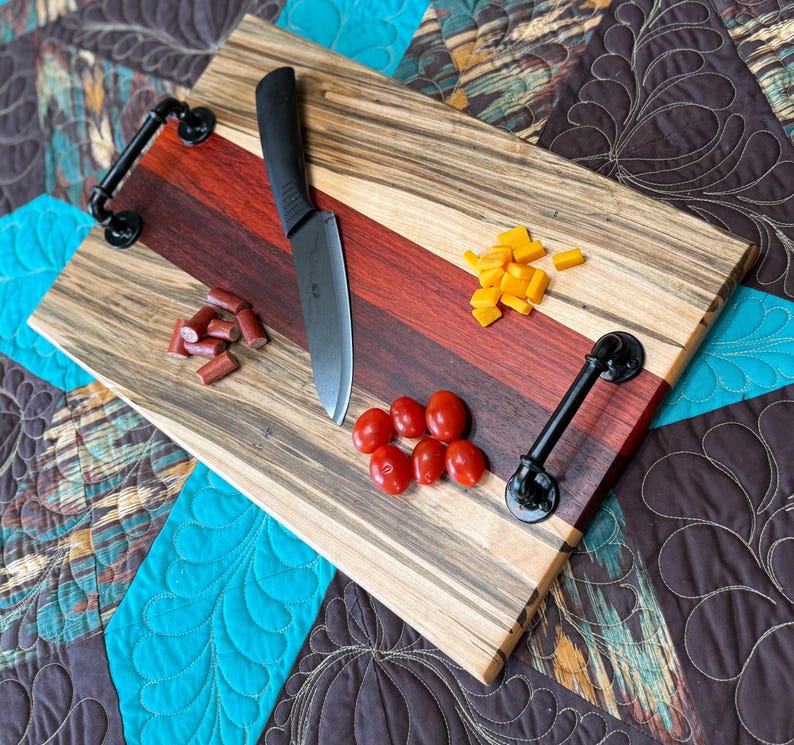 May include: A rectangular wooden serving board featuring alternating stripes of light wood with dark grain and a central deep red wood section. It includes two black metal pipe-style handles, a black kitchen knife, sliced sausage, and cheese.