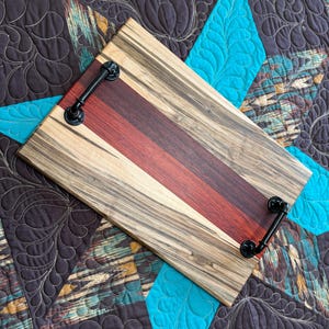 May include: A rectangular wooden serving tray featuring alternating stripes of light tan wood with dark grain patterns and a central band of deep reddish-brown wood. The tray is equipped with two black metal industrial-style handles.