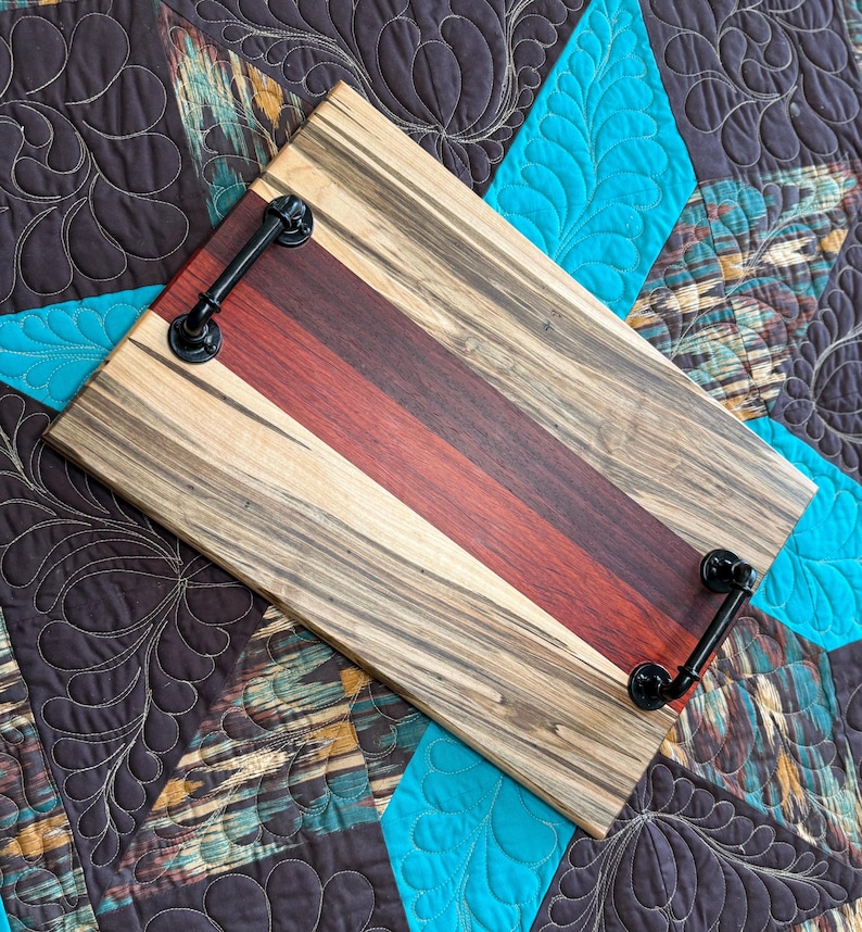 May include: A rectangular wooden serving tray featuring alternating stripes of light tan wood with dark grain patterns and a central band of deep reddish-brown wood. The tray is equipped with two black metal industrial-style handles.