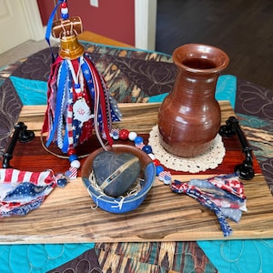 May include: A rectangular wooden serving tray with black metal handles featuring a brown ceramic vase, a small blue bowl containing a dark heart-shaped stone, and a decorative bell ornament adorned with red, white, and blue fabric ribbons.