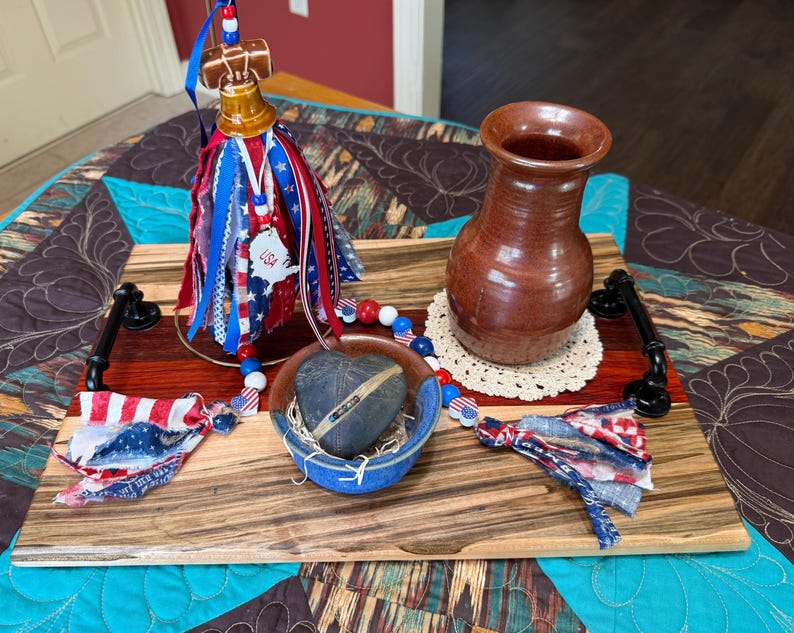 May include: A rectangular wooden serving tray with black metal handles featuring a brown ceramic vase, a small blue bowl containing a dark heart-shaped stone, and a decorative bell ornament adorned with red, white, and blue fabric ribbons.