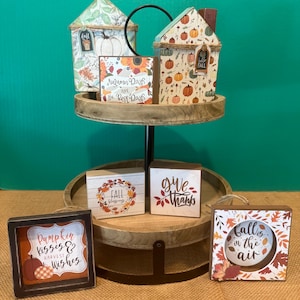 May include: A two-tiered wooden tray holding autumn-themed decor, including two house-shaped signs, three rectangular signs with text like Autumn Days are the Best Days, Give Thanks, and Pumpkin Kisses, and a square sign reading Fall is in the air.