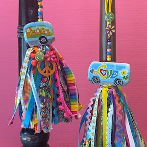 May include: Two decorative hanging ornaments featuring miniature blue camper van charms with the words PEACE and LOVE. Each ornament includes a colorful tassel made of various ribbons, fabric strips, and beads, accented with small flower and smiley face charms.