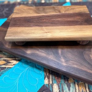 May include: Two wooden serving boards, one stacked on top of the other. The top board is a lighter brown with a square shape and rounded edges. The bottom board is a darker brown with a rectangular shape. The boards are on a patterned quilt.