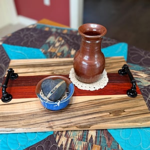 May include: A rectangular wooden serving tray featuring horizontal stripes of light and dark wood grain, equipped with two black metal pipe-style handles. A brown ceramic vase and a small blue bowl containing a dark stone rest on the tray.