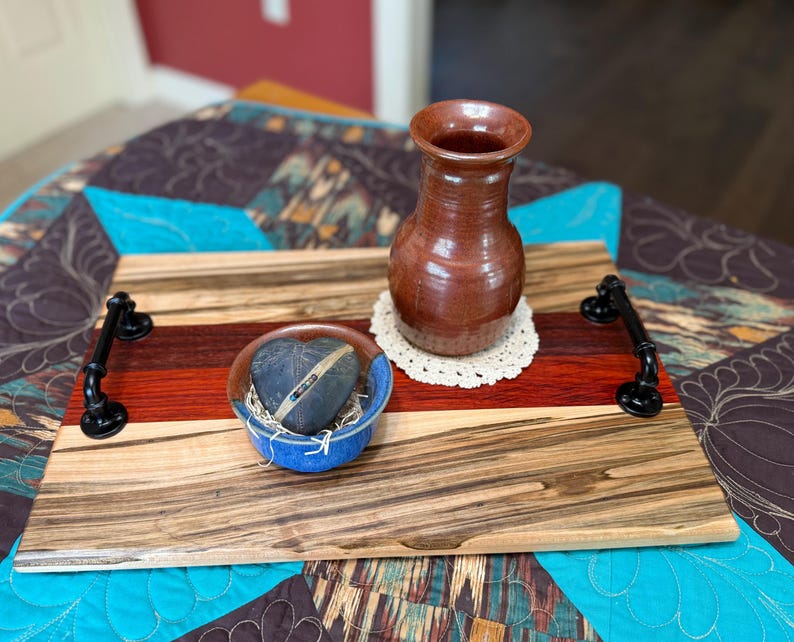 May include: A rectangular wooden serving tray featuring horizontal stripes of light and dark wood grain, equipped with two black metal pipe-style handles. A brown ceramic vase and a small blue bowl containing a dark stone rest on the tray.