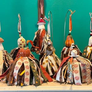 Könnte beinhalten: Eine Sammlung von sechs hängenden Ornamenten mit Keramikfiguren wie Pilzen, Kürbissen, Mais und einem Igel. Jedes Ornament ist mit einem Rock aus verschiedenen Stoffstreifen in herbstlichen Farben, Mustern und Texturen verziert und hängt an Bändern.