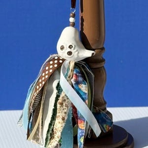 May include: A white ceramic bird ornament with brown eyes and a brown beak hangs from a brown ribbon. The bird is adorned with a colorful fabric tassel featuring various patterned ribbons in shades of blue, brown, and white.