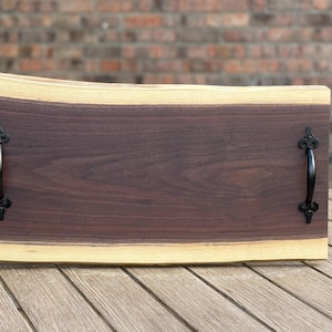 May include: A rectangular wooden serving tray with a dark brown center and a light brown edge. The tray has black metal handles on each end. The wood grain is visible. The tray is on a wooden table.