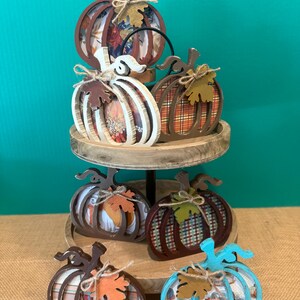 May include: A two-tiered wooden tray holding seven decorative pumpkin cutouts. Each pumpkin features a unique cutout frame, a patterned insert, and a small wooden leaf accent tied with twine. The pumpkins vary in colour, including brown, white, and teal.