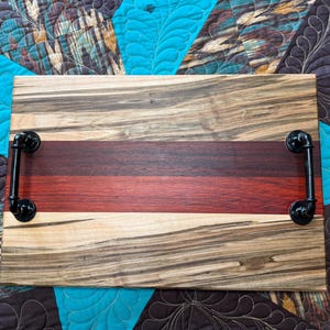May include: A rectangular wooden serving tray featuring a multi-toned wood grain pattern with a prominent dark red horizontal stripe through the center. The tray is equipped with two black industrial-style metal pipe handles on each side.