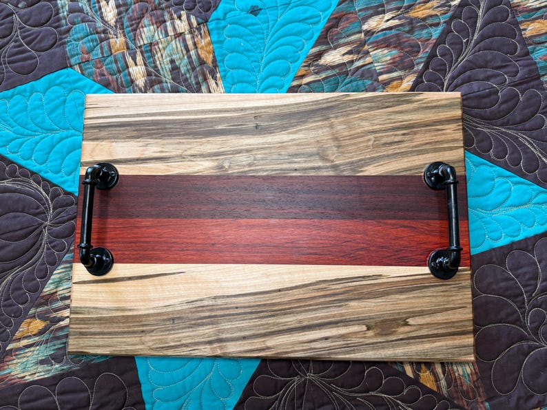 May include: A rectangular wooden serving tray featuring a multi-toned wood grain pattern with a prominent dark red horizontal stripe through the centre. The tray is equipped with two black industrial-style metal pipe handles on each side.