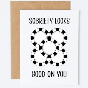May include: A white greeting card with the text "SOBRIETY LOOKS GOOD ON YOU" in black. The card features a graphic of four overlapping poker chips with black and white stripes. The card is paired with a brown envelope.