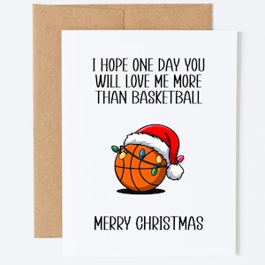 Op de afbeelding: Een witte wenskaart met een bruine envelop met de tekst I HOPE ONE DAY YOU WILL LOVE ME MORE THAN BASKETBALL en MERRY CHRISTMAS. De afbeelding in het midden toont een oranje basketbal met een kerstmuts en een snoer met kerstverlichting.