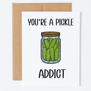 Peut inclure: Carte de vœux blanche avec le texte « YOU'RE A PICKLE ADDICT » et une illustration d'un bocal rempli de cornichons verts. La carte est posée sur une enveloppe marron.