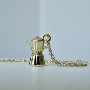 May include: A gold pendant necklace with a miniature coffee maker charm. The charm has a decorative band of clear crystals. The necklace chain and charm are made of shiny, gold-toned metal. The coffee maker charm is approximately 2.5 cm tall.