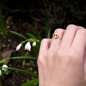 May include: A gold ring with a heart-shaped design featuring a small pink stone in the centre. The ring is worn on a hand holding a sprig of white flowers.