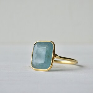 Könnte beinhalten: Ein goldener Ring mit einem großen, hellblauen Aquamarin-Edelstein, der in einer quadratischen Form gefasst ist.