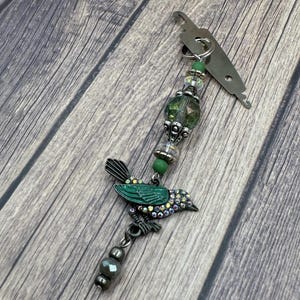 May include: A green bird charm with rhinestones and a green glass bead hangs from a silver threader. The threader has a metal loop at the top.