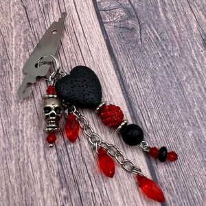 May include: A black lava rock heart charm with red crystal beads, a silver skull charm, and a silver chain. The charm is attached to a silver hook.