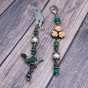 May include: Two decorative keychains with beaded accents. One features a green bird charm with rhinestone accents, the other has peach-colored rose charms. Both have silver-tone hardware and a mix of green, gray, and pearl-like beads.