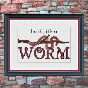May include: Framed cross-stitch art with the text "Look, it's a WORM." The artwork features a brown worm design and text in a dark burgundy color. The frame is black with a burgundy inner border. The background is a brick wall.