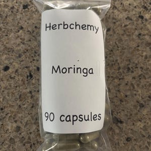May include: A clear plastic bag containing 90 capsules. A white label on the bag reads "Herbchemy Moringa" in black text. The capsules are a light green color.