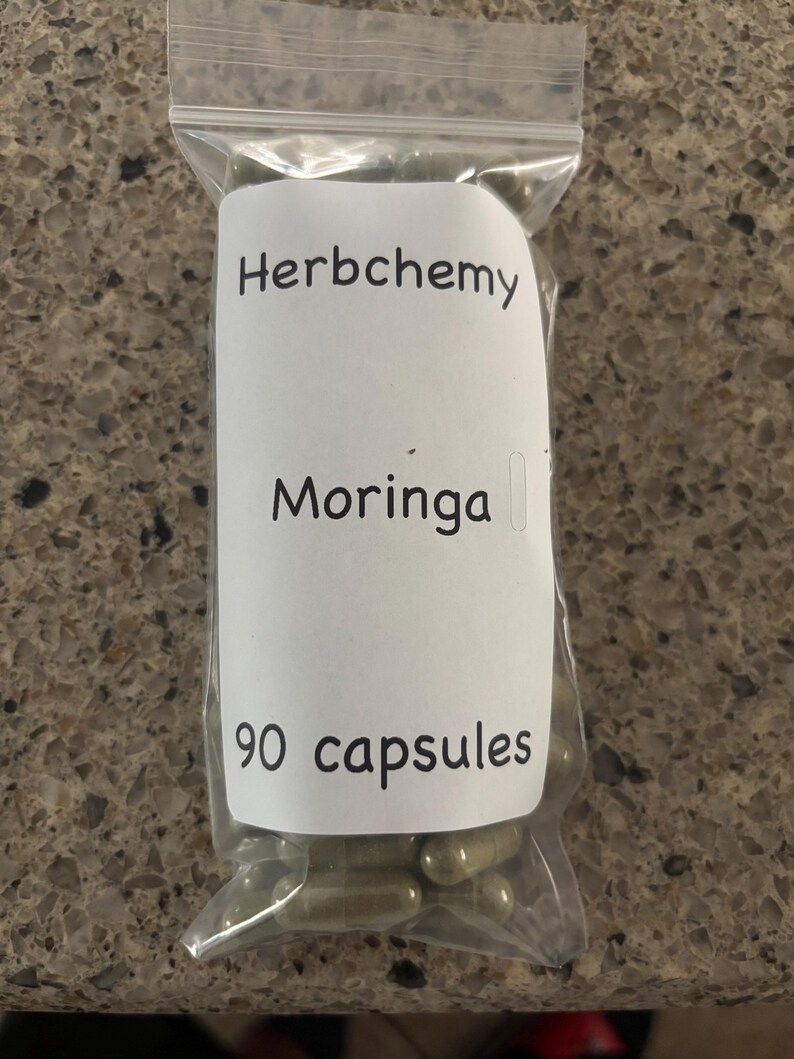 May include: A clear plastic bag containing 90 capsules. A white label on the bag reads "Herbchemy Moringa" in black text. The capsules are a light green color.