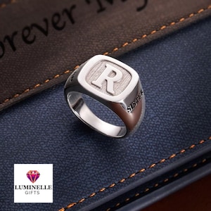 Könnte beinhalten: Ein silberner Siegelring mit dem Buchstaben "R" in einem quadratischen Rahmen. Das Wort "SISTER" ist seitlich eingraviert. Der Ring wird auf einer blauen Oberfläche mit einem braunen Lederhintergrund präsentiert.
