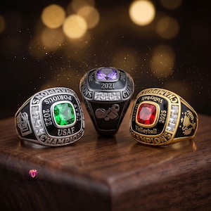 Könnte beinhalten: Drei Meisterschaftsringe, bestehend aus einem silbernen Ring mit grünem Stein, einem schwarzen Ring mit violettem Stein und einem goldenen Ring mit rotem Stein. Jeder Ring zeigt Inschriften wie "USAF 2022", "Duke 2021" und "Volleyball 2025".