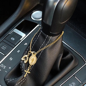 Op de afbeelding: Gouden auto-hanger met een hart, een ovale hanger met een religieuze figuur en een kruis. De hanger hangt aan een gouden ketting en wordt getoond op een zwarte versnellingspook.