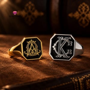 May include: Two signet rings with octagonal faces and black enamel inlays. The left ring is gold-toned with a gold letter A monogram. The right ring is silver-toned with a silver letter K monogram. Both feature intricate, decorative line work.