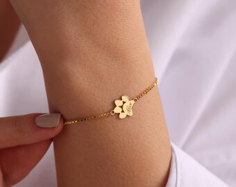 Puede incluir: Una pulsera dorada con una cadena delicada. La pulsera presenta un dije en forma de pata con el nombre "Alex" grabado. La pulsera se lleva en la muñeca.