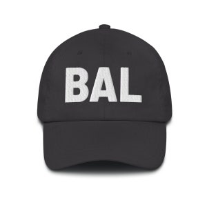 May include: A dark gray baseball cap with the embroidered letters "BAL" in white across the front. The cap has a curved brim and a fabric strap closure on the back. The hat is a classic style.