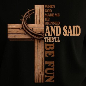 Puede incluir: Camiseta negra con un diseño de cruz de madera marrón. La cruz tiene una corona de espinas y el texto "When God made me he grinned and said this'll be fun".