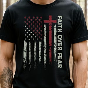 Könnte beinhalten: Schwarzes T-Shirt mit einem verwitterten Design der amerikanischen Flagge. Das Design beinhaltet ein rotes Kreuz und die Worte "FAITH OVER FEAR" in Weiß. Das Flaggen-Design enthält Kreuze und Sterne.