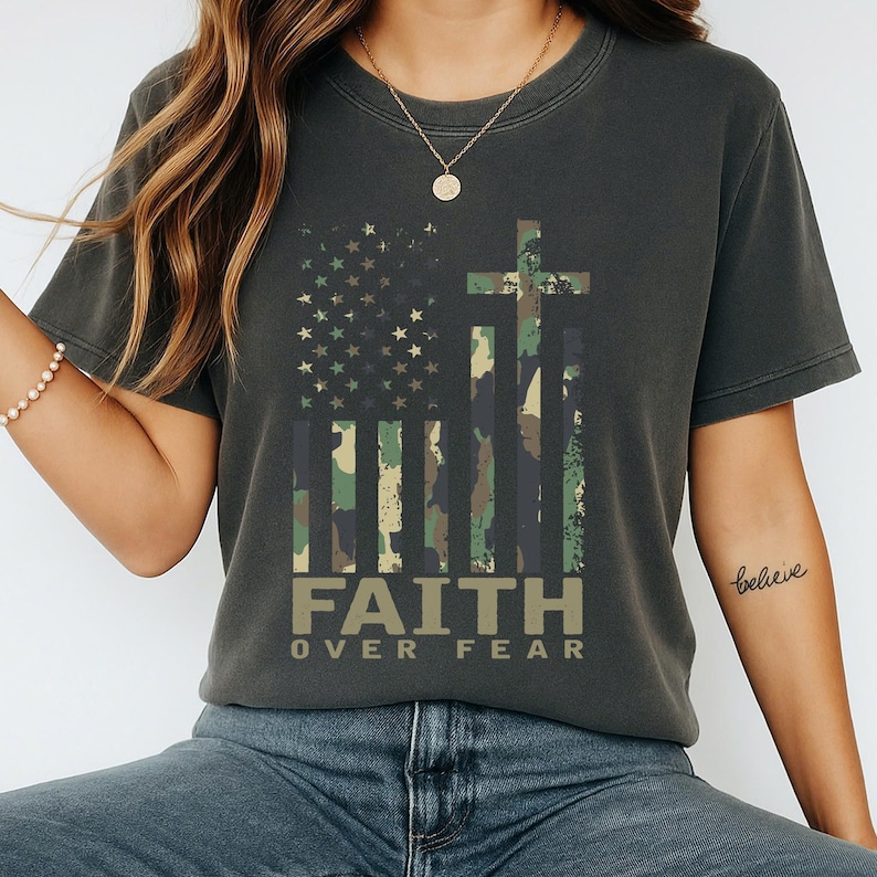 May include: Dark gray t-shirt featuring a camouflage American flag design with a cross and the words "FAITH OVER FEAR" in a tan font. The shirt has a crew neck and short sleeves, suitable for casual wear.
