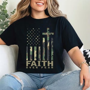 May include: Black t-shirt featuring a camouflage American flag design with a cross and the words "FAITH OVER FEAR" in a bold font. The shirt has short sleeves and a relaxed fit, suitable for casual wear.