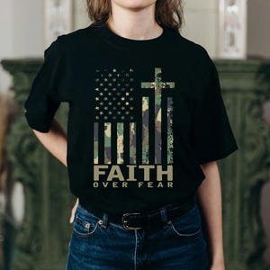 May include: Black t-shirt featuring a camouflage American flag design with a cross and the words "FAITH OVER FEAR" in a bold, sans-serif font. The flag design incorporates stars and stripes in a distressed style, creating a vintage look.