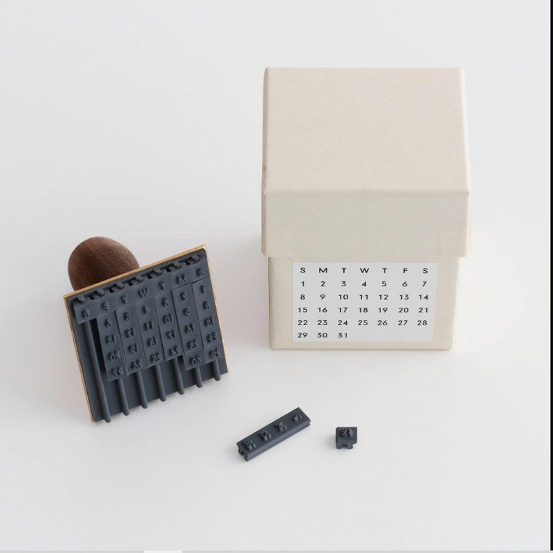 May include: A set of gray rubber stamps in a box. The stamps are for a calendar and include the days of the week and numbers 1 through 31. The box is beige with black text.