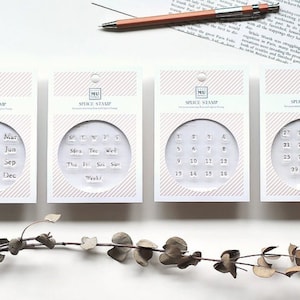 May include: Four rectangular packages of MU Splice Stamp sets with pink striped headers. Each package contains clear, square rubber stamps featuring months, days of the week, and numbers 1 through 31 for calendar marking.