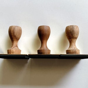 May include: Three wooden stamp blocks with a dark brown finish. Each block has a rectangular base and a rounded top. The blocks are arranged in a row on a black surface.