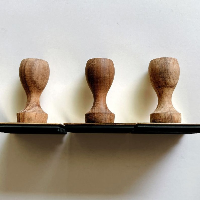 May include: Three wooden stamp blocks with a dark brown finish. Each block has a rectangular base and a rounded top. The blocks are arranged in a row on a black surface.